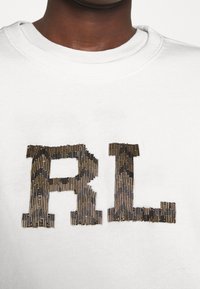 Bílá bavlněná mikina s texturovaným logem "RL" v metalické zlaté a hnědé pruhy. Hladký výstřih a krátké rukávy.