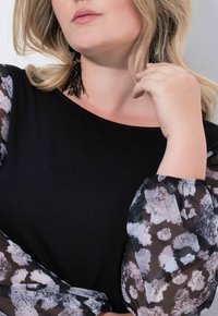 Czarny top z przezroczystymi, wzorzystymi rękawami w szaro-białym wzorze. Rękawy są marszczone przy mankietach. Widoczne czarne kolczyki na wisząco.