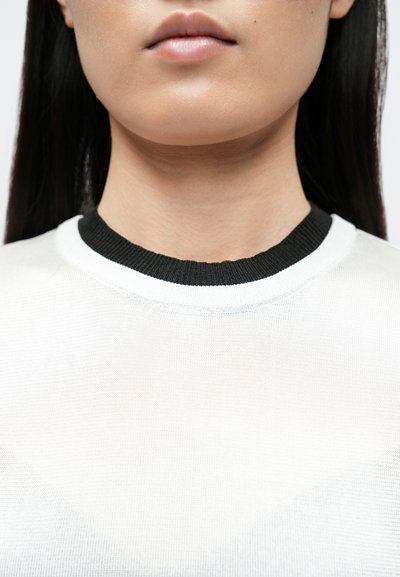 Haut en maille blanche avec un col côtelé noir, présentant une texture lisse et un design simple. Détails de coutures visibles au niveau de l'encolure.