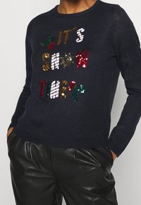 Pull en tricot bleu marine avec du texte en sequins multicolores "C'EST L'HEURE DE LA NEIGE" et des accents de houx verts. Col et poignets côtelés.