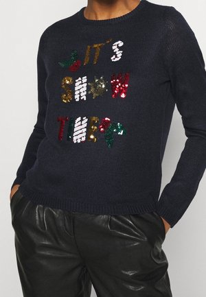 Pull en tricot bleu marine avec du texte en sequins multicolores "C'EST L'HEURE DE LA NEIGE" et des accents de houx verts. Col et poignets côtelés.