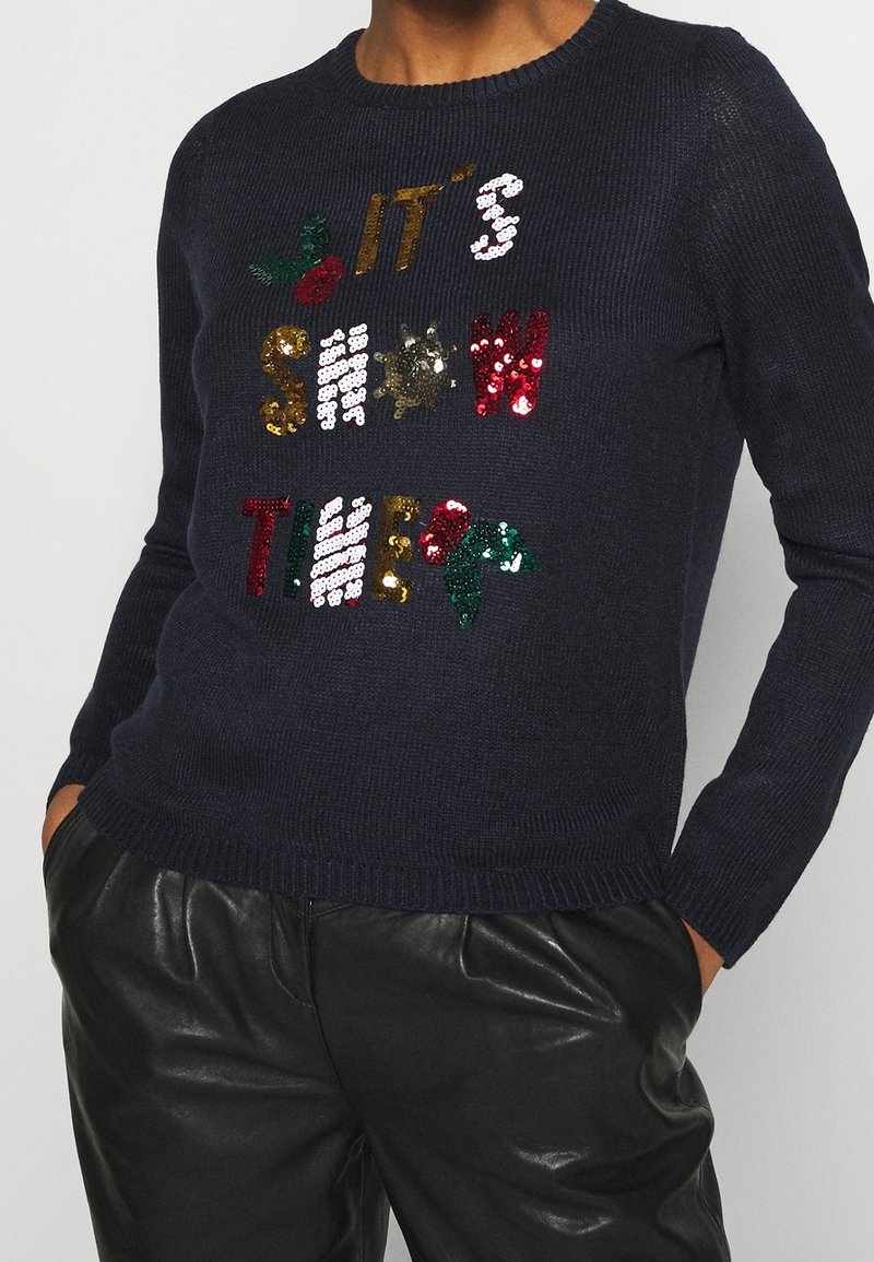 Pull en tricot bleu marine avec du texte en sequins multicolores "C'EST L'HEURE DE LA NEIGE" et des accents de houx verts. Col et poignets côtelés.