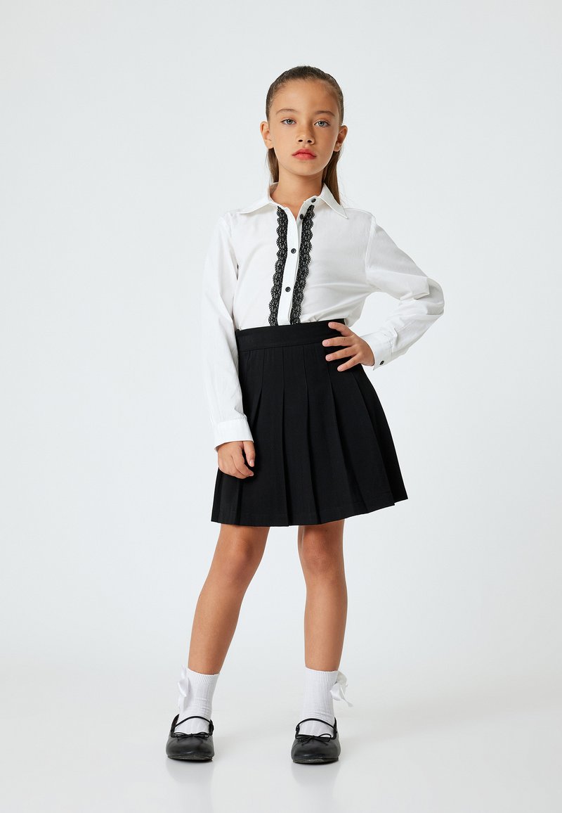 Camicia bianca con rifiniture in pizzo nero, gonna plissettata nera, calze bianche e scarpe nere con fiocchi. Tessuto liscio, design semplice.
