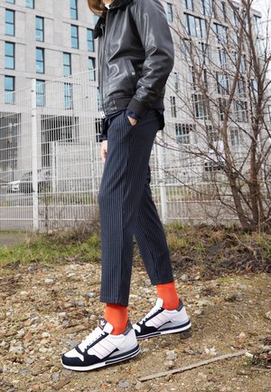 Persoon staand op rotsachtig terrein, gekleed in een zwart leren jack, pantalon met krijtstrepen, feloranje sokken en zwart-witte sneakers.