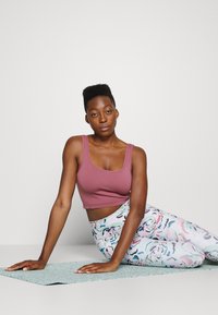 Top cropped de cor mauve com decote em forma de U e textura canelada, combinado com leggings multicoloridas com um padrão floral. Pessoa em um tapete de yoga texturizado.