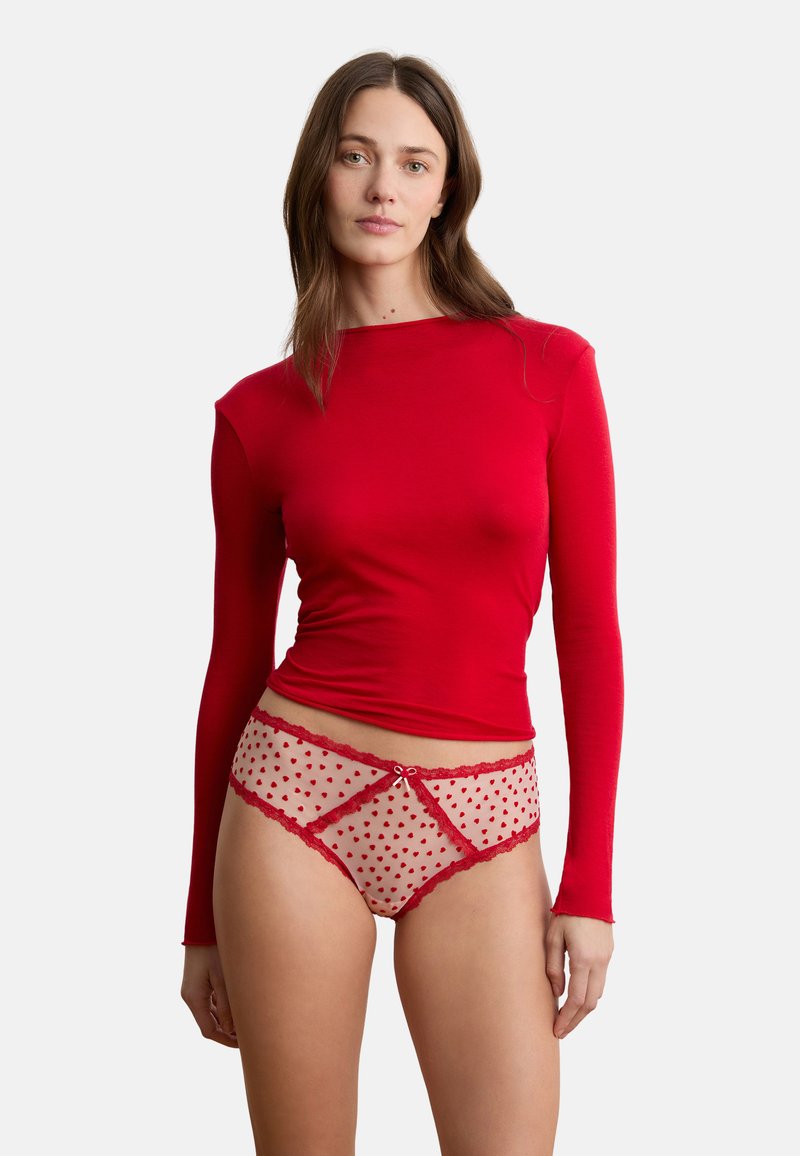 Vrouw die een lange mouw rode top draagt en doorschijnende beige slip met rode hartpatronen en kanten details, staand tegen een witte achtergrond.