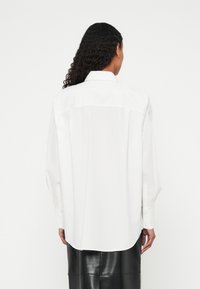 Ženska z dolgimi kodrastimi lasmi, oblečena v ohlapno belo majico z dolgimi rokavi in črno usnjeno krilo, prikazana od zadaj na enobarvni podlagi.