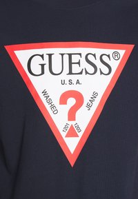 T-shirt in cotone blu navy con una grande grafica triangolare rossa che presenta il logo "GUESS", il testo "WASHED JEANS" e un punto interrogativo centrale.