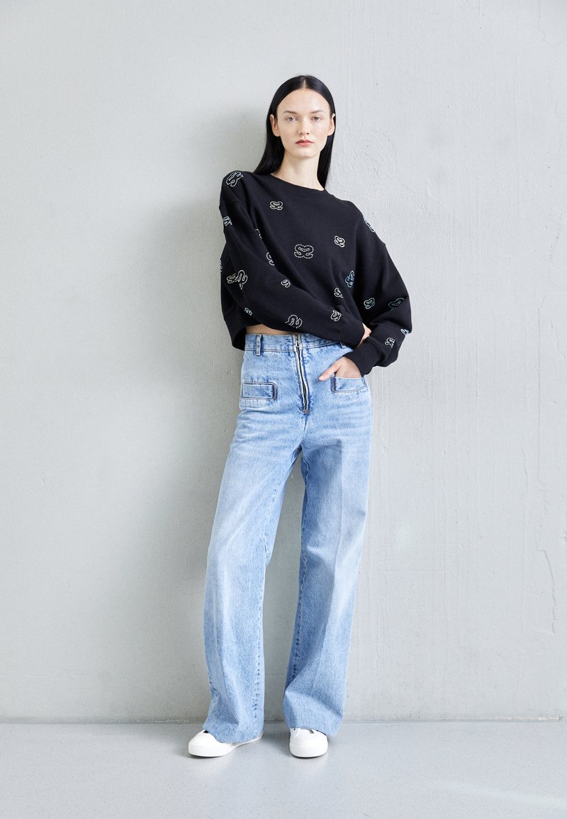Schwarzer Pullover mit bunten abstrakten Mustern, kombiniert mit hellblauen weit geschnittenen Jeans und weißen Sneakers. Insgesamt glatte Textur.