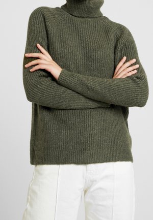 Person trägt einen gerippten olivgrünen Rollkragenpullover mit verschränkten Armen, kombiniert mit lockeren weißen Hosen vor einem einfarbigen Hintergrund.