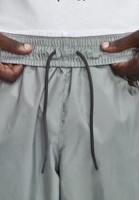 Calções cinza claro com cintura elástica, cordão preto e textura suave ligeiramente brilhante. Detalhes incluem dois bolsos laterais.