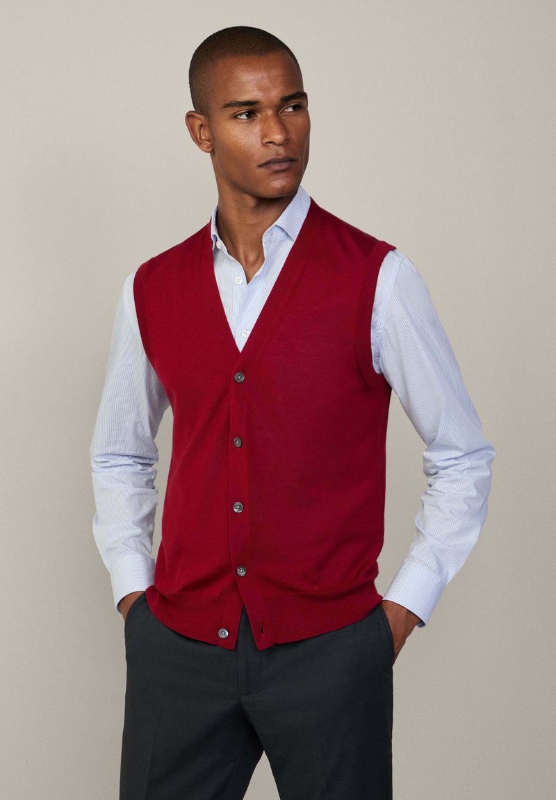 Gilet rosso a maglia con scollo a V, chiusura con bottoni e senza maniche, indossato sopra una camicia a righe azzurro chiaro con colletto. Pantaloni grigi.