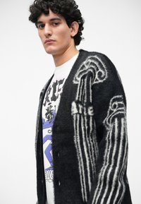 Cárdigan de punto en blanco y negro con textura suave, patrones de rayas en las mangas y cierre de botones, llevado sobre una camiseta gráfica.