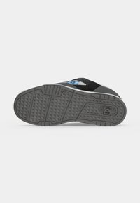 Semelle de chaussure vue de dessous avec une bande de roulement grise texturée et un logo bleu près du talon sur une basket noire et grise.