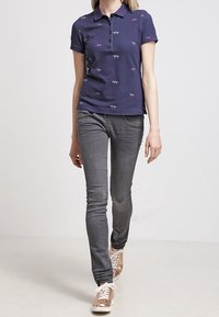 Marinblå polo med kort ärm och diskret djurmönster, i kombination med slim-fit grå jeans och metalliska sneakers för en avslappnad stil.