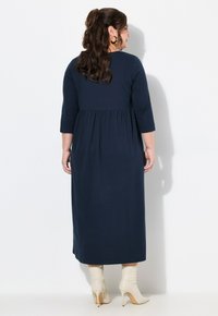 Marineblauwe maxi-jurk met driekwartmouwen, een uitlopende onderkant en een geplooide taille. Gedragen met witte haklaarzen.