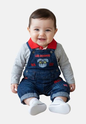 Bébé souriant portant une chemise gris à manches longues, un col rouge, une salopette en denim avec un motif de chien, et des chaussures blanches à semelle souple, assis sur un fond blanc.