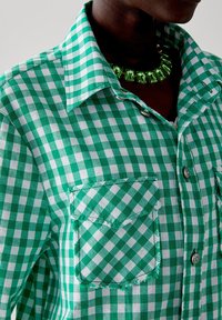 Camicia a quadri verde e bianca con colletto classico, taschino sul petto e bottoni argentati. Il tessuto sembra texture e leggero.