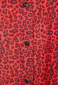 Rød stof med et leopardprint mønster i mørkere nuancer, med sorte knapdetaljer foran. Glat tekstur, skræddersyet pasform.