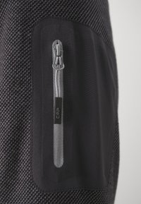 Tissu noir avec un motif texturé, présentant une poche zippée avec une fermeture éclair grise et le logo CMP. Matériau lisse et durable.