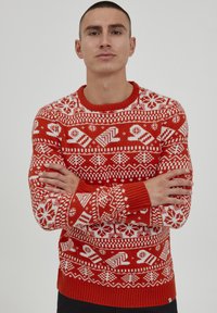Pull en tricot rouge avec des motifs d'hiver blancs, incluant des flocons de neige et des designs géométriques, avec un col rond et des poignets côtelés.