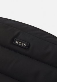 Černá polstrovaná tkanina se stříbrným štítkem s logem "BOSS". Zip je umístěn podél horního okraje a má hladkou texturu.