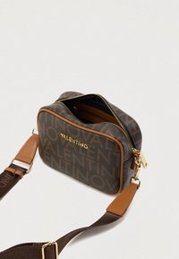 Brązowa torba na ramię z tkaniny z powtarzającym się wzorem logo, złotymi elementami, zamkiem błyskawicznym i odpinanym paskiem z skórzanymi akcentami.
