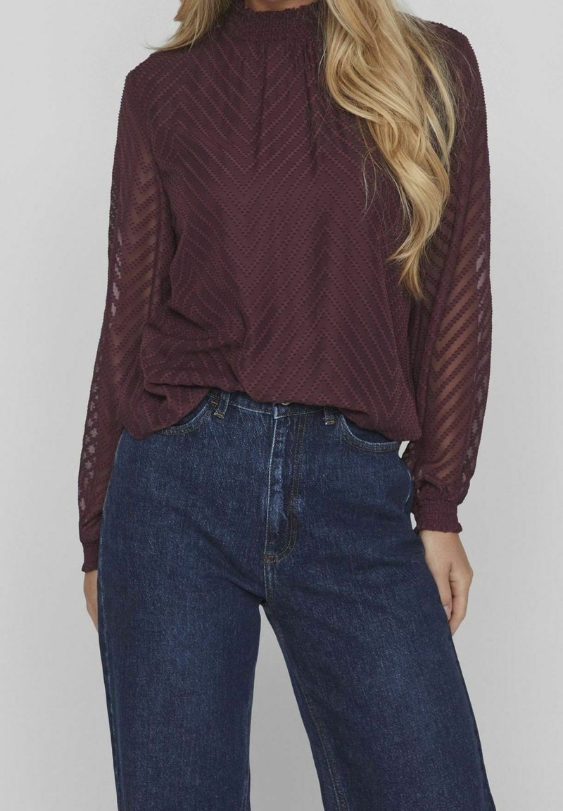 Femme portant un chemisier bordeaux transparent et texturé à manches longues, rentré dans un jean en denim bleu foncé taille haute, sur un fond uni.