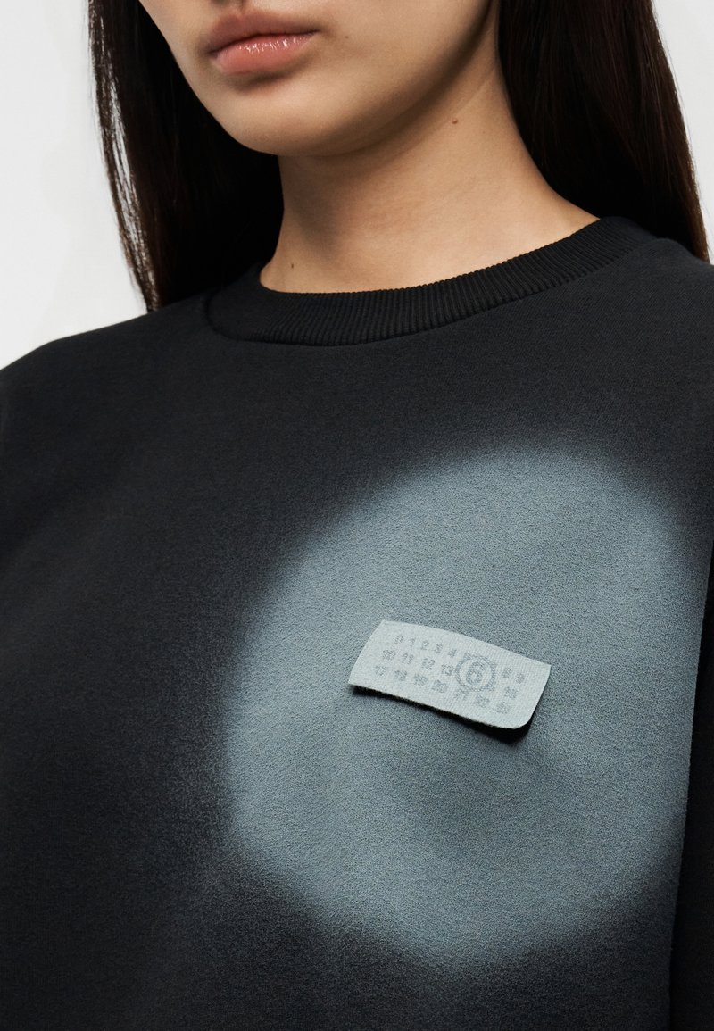 Sweat-shirt gris foncé avec un col arrondi. Présente une étiquette gris clair sur la zone de la poitrine avec un motif texturé et un fond à dégradé subtil.