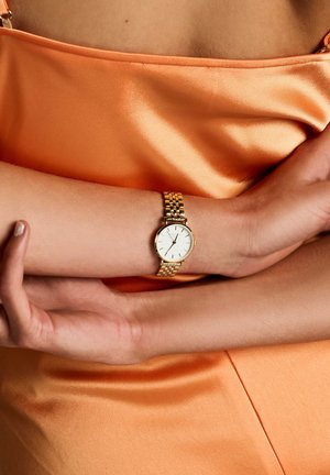 Gold wristwatch with white face, gold metal band, simple round case, worn on a wrist against a satin orange fabric background.