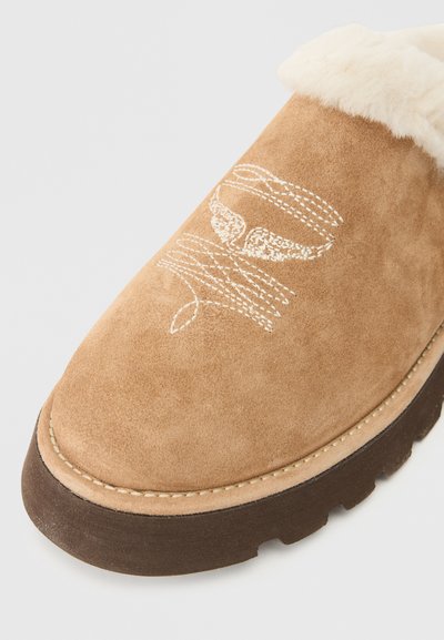 Sapato slip-on de camurça em castanho claro com colarinho de pêlo sintético, apresentando costura decorativa branca e uma sola de borracha texturizada para melhor aderência.