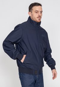 Marineblaues Bomberjacke mit hohem Kragen, Reißverschluss und elastischen Bündchen. Verfügt über Seitentaschen und unauffällige Markenkennung auf der Brust. Glatte Textur.