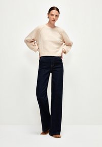 Beiger, verkürzter Pullover mit weiten Ärmeln, kombiniert mit dunkellblauen High-Waist-Jeans. Glatte Textur, keine sichtbaren Muster, ergänzt durch tanfarbene Schuhe.