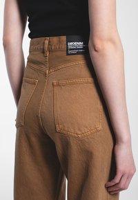 Pantaloni in denim marrone con vita alta, tasche posteriori e un'etichetta nera in vita. Il tessuto ha una finitura strutturata.