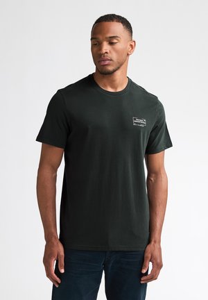 Mann trägt ein schlichtes schwarzes Kurzarm-T-Shirt mit kleinem weißem "Petrol Industries"-Logo auf der linken Brust, stehend vor weißem Hintergrund.
