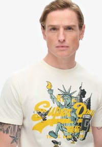 Camiseta de algodón color crema con un diseño gráfico de la Estatua de la Libertad, rascacielos y pájaros en acentos amarillos y negros.