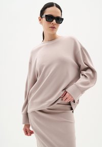 Oversized ljusrosa sweatshirt med rund halsringning och långa ärmar, tillsammans med en matchande midikjol. Modellen bär svarta solglasögon.