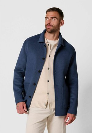 Man met kort haar en baard, draagt een blauwe gecoate jas over een beige vest en lichtgekleurde broek, kijkt zijwaarts.
