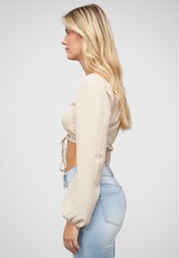 Top beige a maniche lunghe con finitura texturizzata e dettaglio con fiocco frontale, abbinato a jeans azzurri a vita alta, visto di lato.