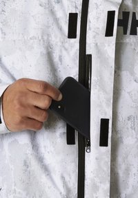 Bílá bunda s texturovým povrchem, se zipovou kapsou. Ruka vkládá černý telefon do kapsy, což zdůrazňuje funkční design.