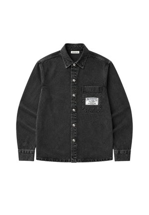Mann mit schwarzer Kappe, ausgewaschener schwarzer Jeansjacke mit Brusttaschen-Applikation, hellblauem Reißverschluss-Pullover und schwarzen Nadelstreifenhosen.