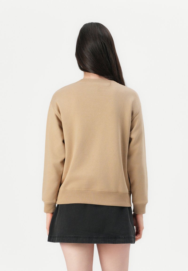 Mujer con pelo largo y oscuro, vistiendo una sudadera beige y una falda negra, de pie de espaldas contra un fondo blanco liso.
