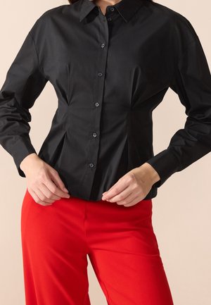 Kobieta w dopasowanej czarnej bluzce zapinanej na guziki z długimi rękawami i szpiczastym kołnierzykiem, w zestawieniu z wysokim stanem i jasnoczerwonymi spodniami.