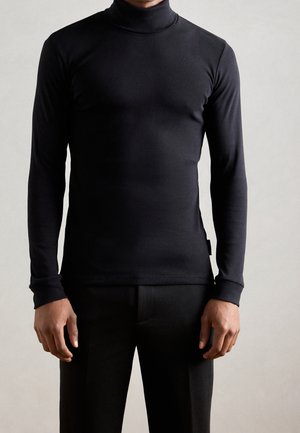 Personne portant un col roulé noir ajusté à manches longues et un pantalon noir, debout devant un fond clair uni.