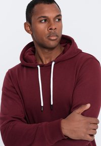 Felpa bordeaux realizzata in morbido tessuto con cappuccio regolabile, dettagli bianchi e polsini a costine. Presenta una vestibilità regolare e un design minimal.