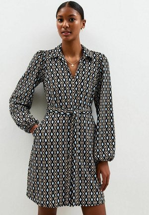 Femme portant une robe à manches longues cintrée avec un motif en diamants noir, blanc et bleu clair, se tenant devant un fond gris clair uni.