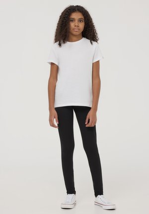 T-shirt bianco in cotone con maniche corte, abbinato a leggings neri. Modella che indossa sneakers bianche, in piedi su uno sfondo chiaro.