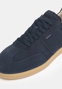 Zapatilla de ante azul marino con punta redondeada, cordones en la parte delantera y suela de goma marrón claro. Presenta un sutil logotipo de la marca en el lateral.