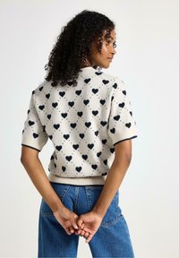 Kortærmet sweater i lys beige med sort hjerte-mønster, ribbede manchetter og søm. Bæres med blå højtaljede jeans.