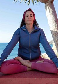 Felpa blu con zip, leggings rosa con motivo testurizzato, posa seduta vicino al mare. Capelli lunghi e scuri, fascia per capelli, espressione calma. Palma visibile.